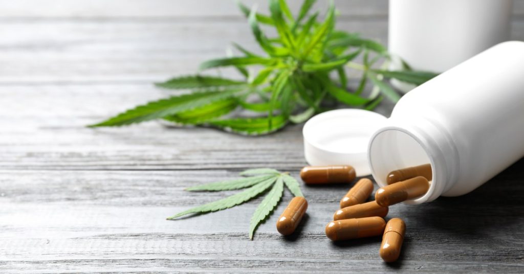 Cannabis leaves next to a white bottle lying down on a table. The bottle has brown capsules spilling out of it.