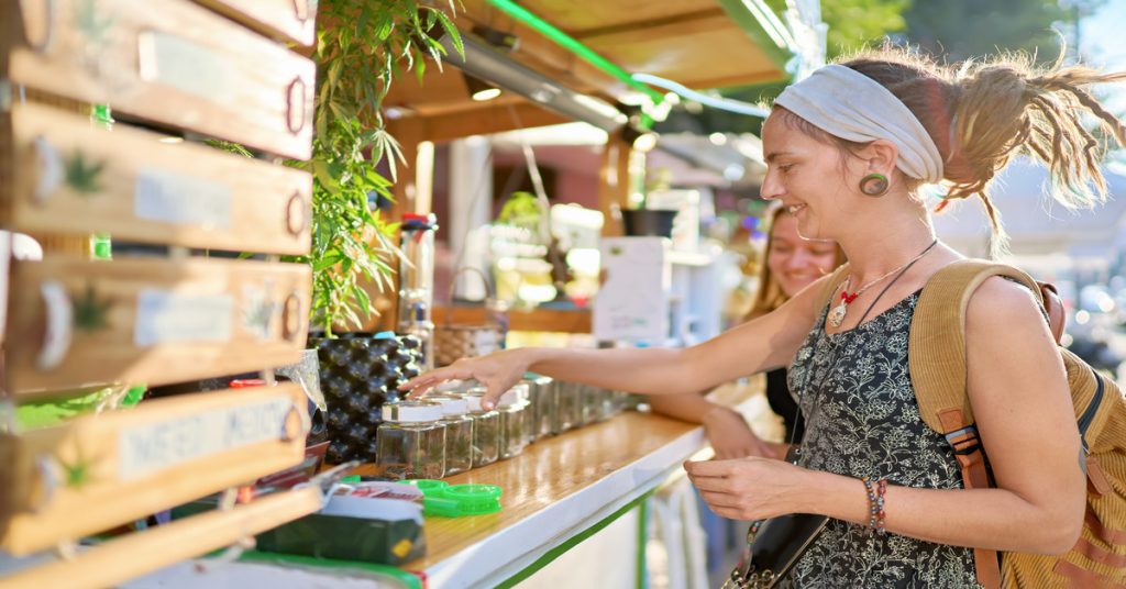Two women examine various cannabis strains at a stand. One woman wearing a backpack grabs a jar and smiles.