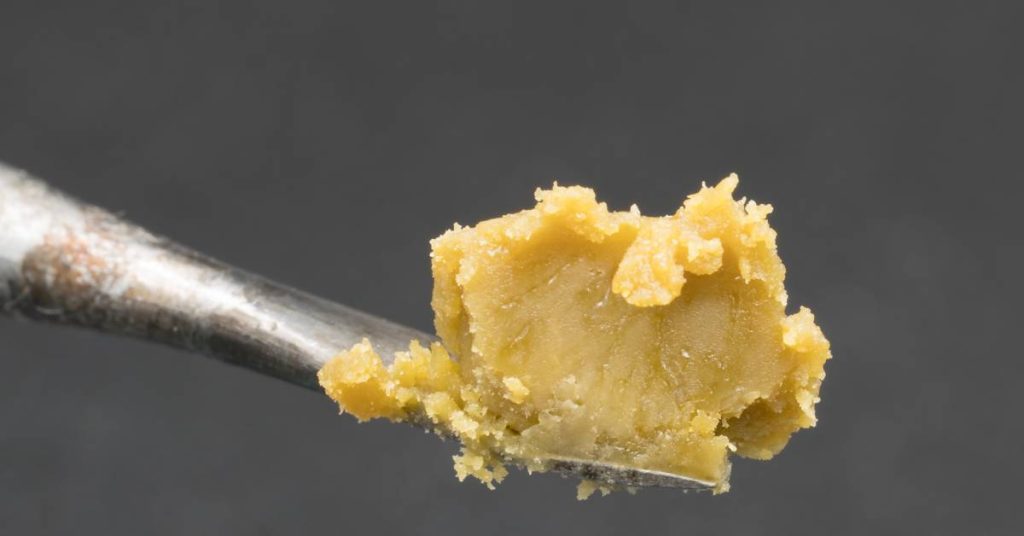 Close-up of rosin on a metal tool in front of a gray background. The rosin has a muted yellow, shea-butter-like color.