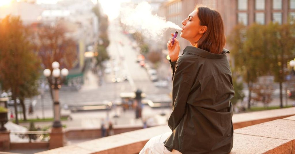 A woman wears a dark grey shirt and sits on a stone bench while vaping. She's facing a blurred city.