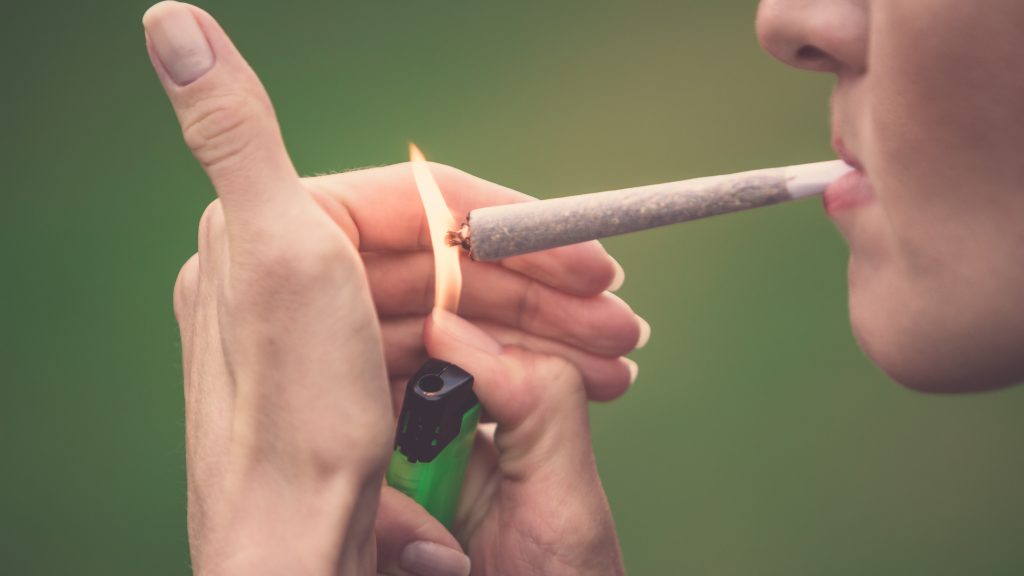 A woman lighting a cannabis joint in front of a green background. She cups her hand around the flame.