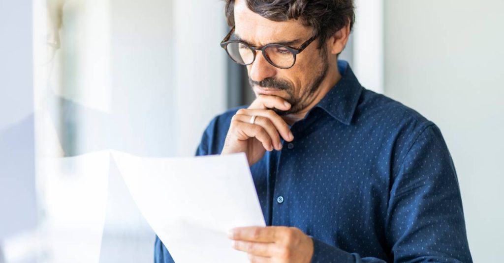  A man wearing black glasses is reading a document intently, his finger to his chin. He’s also standing near a window.